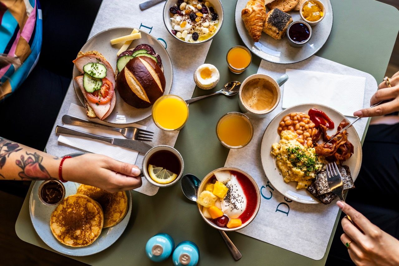Verschiedene Frühstücksgerichte auf Tisch: Pancakes, belegte Brötchen, Obst- und Joghurtbowls, Rührei mit Bohnen, Croissants, Kaffee, Orangensaft und Tee. Zwei Personen greifen nach Essen, gemütliche Atmosphäre.
