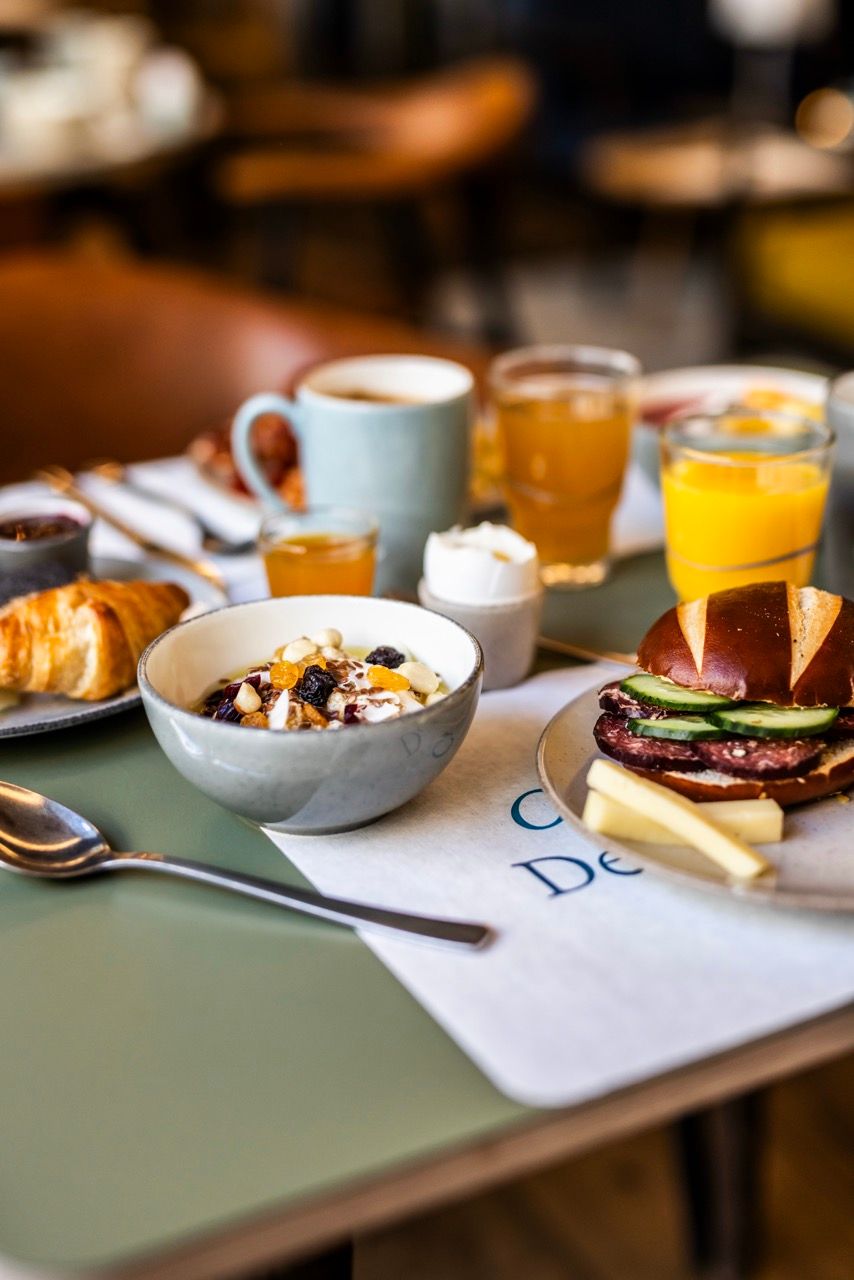 Frühstückstisch mit Joghurt und Müsli, Croissant, Brezel-Sandwich mit Salat und Fleisch, Kaffee, Orangensaft und weiteren Getränken, in gemütlicher Atmosphäre.