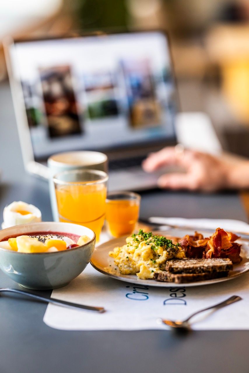 Frühstückstablett mit Rührei, Speck, Brot, Obst und Säften vor einem Laptop, das eine entspannte Morgenatmosphäre für Geschäftsreisende oder Urlauber vermittelt.