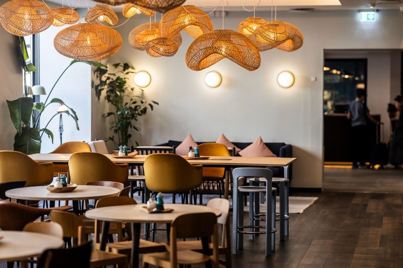 Gemütlicher Restaurantbereich mit Holztischen, bequemen Stühlen und sanfter Beleuchtung durch dekorative Flechtlampen, umgeben von Pflanzen und ruhiger Atmosphäre für entspannte Mahlzeiten oder Gespräche.