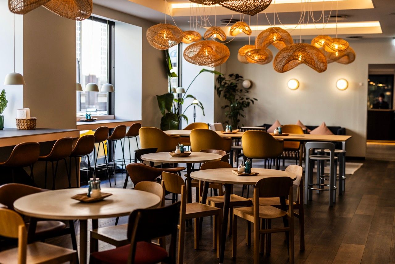 Moderne Hotel-Lounge mit warmem Licht, runden Holztischen und bequemen Stühlen, dekoriert mit Pflanzen und einzigartigen Flechtlampen, ideal für entspannte Mahlzeiten oder Gespräche in stilvoller Atmosphäre.