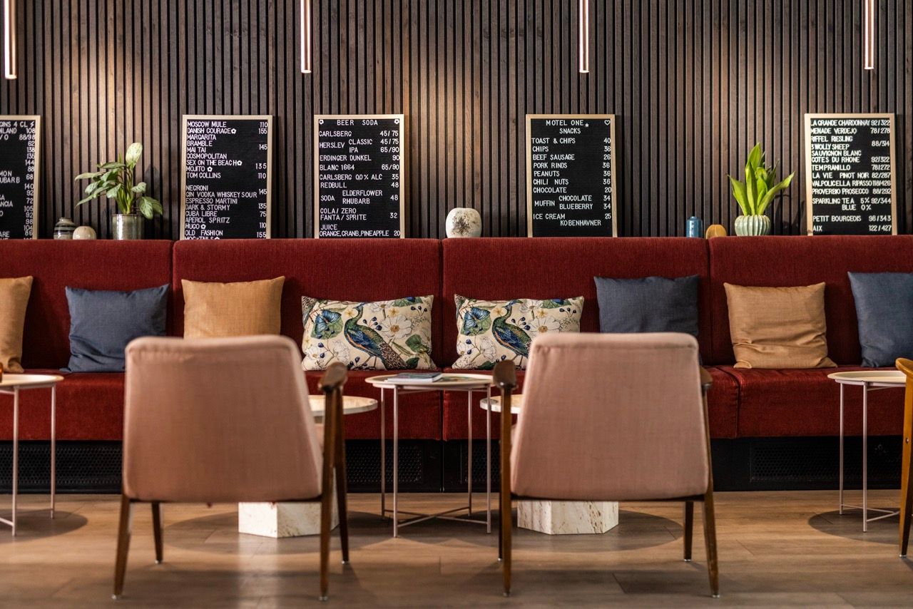 Modernes Hotel-Lounge mit rotem Sofa, bunten Kissen und bequemen Stühlen. Schwarze Speisekarten an der Wand zeigen Getränke- und Snackauswahl. Stilvolle Atmosphäre für entspannte Pausen oder informelle Treffen.