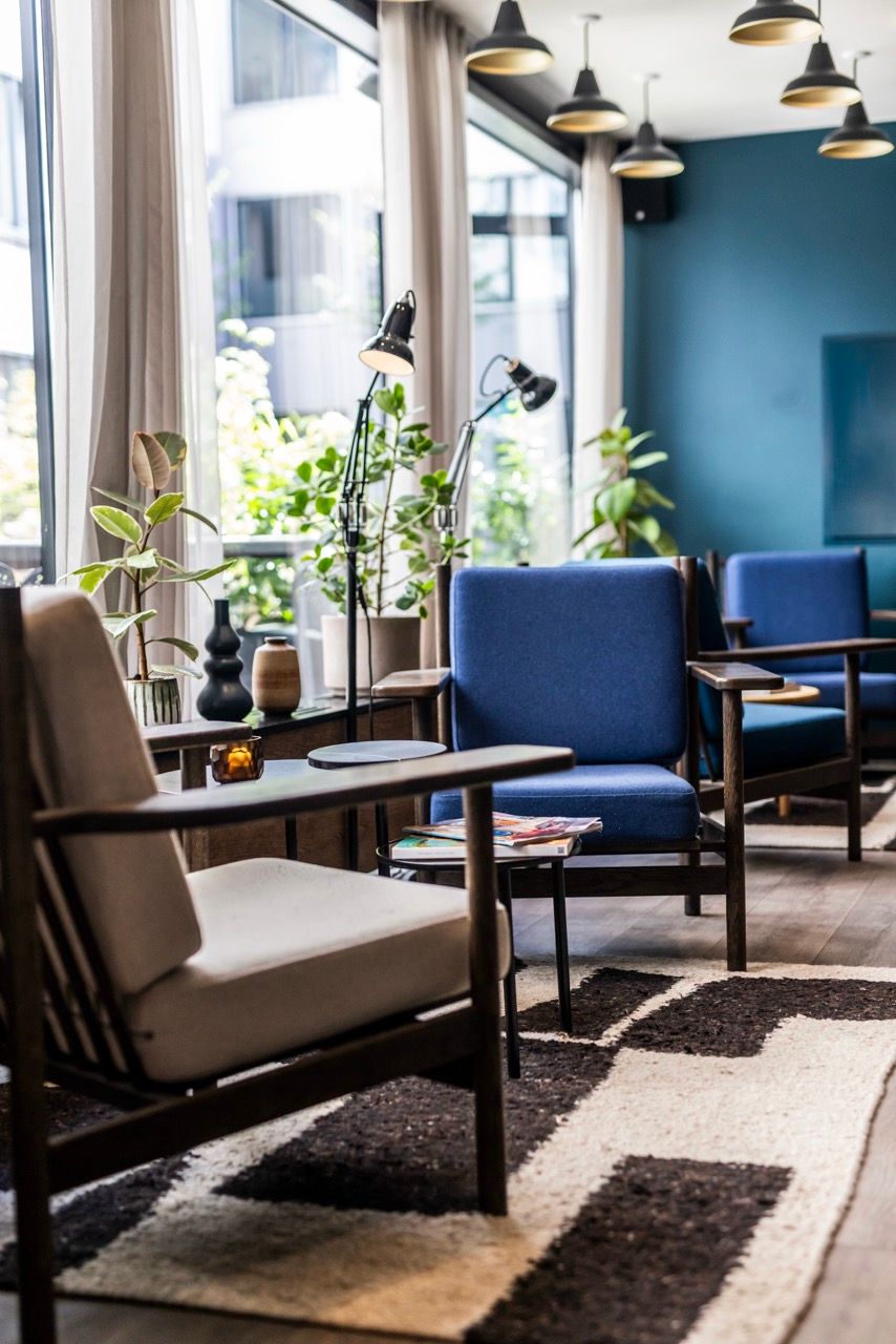 Gemütlicher Sitzbereich mit bequemen Sesseln in Blau und Beige, modernem Lichtkonzept und großen Fenstern, die viel Tageslicht und grüne Pflanzen hereinlassen. Wohnzimmeratmosphäre für entspannte Momente oder geselliges Beisammensein.