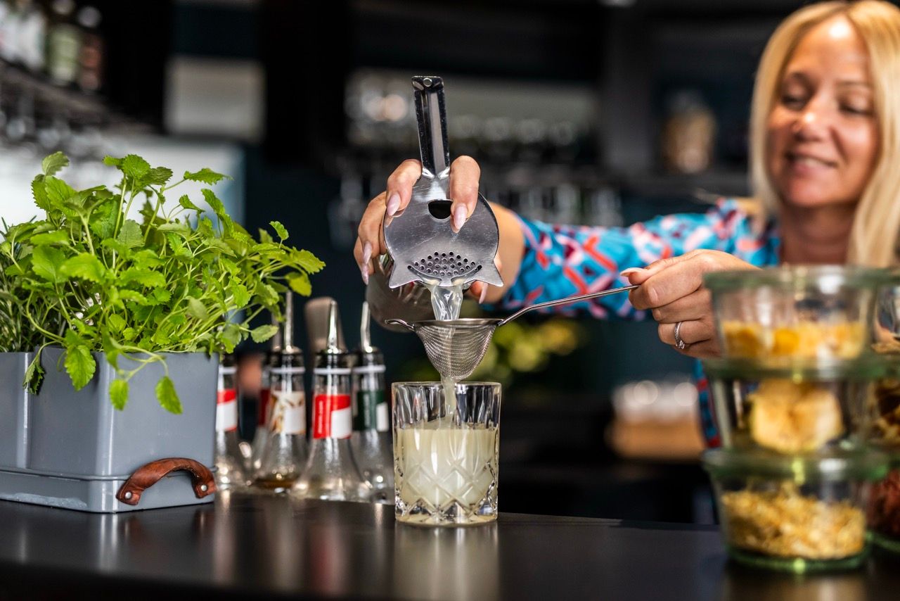 Eine Barkeeperin siebt einen Cocktail in ein Glas, umgeben von frischen Kräutern und Barutensilien, in einer modernen Bar.
