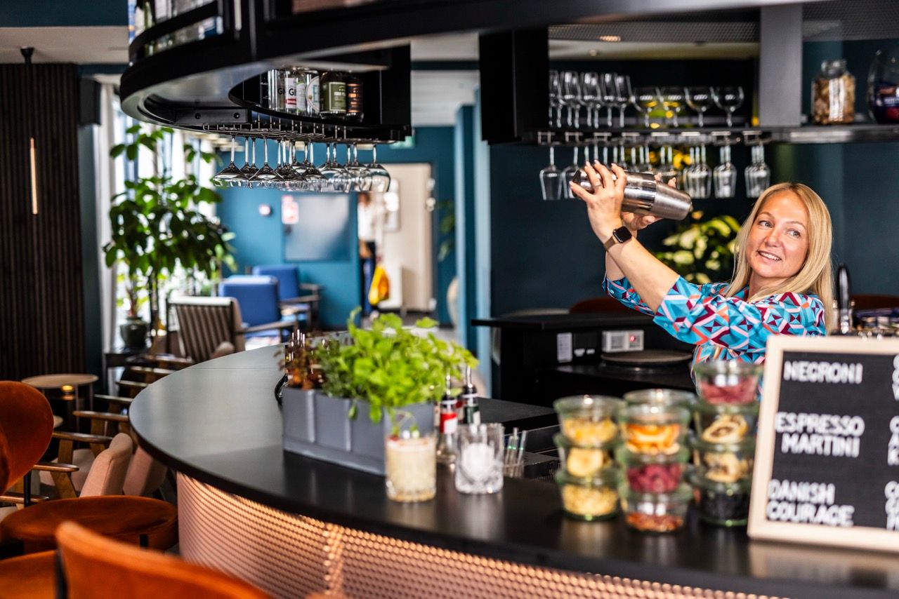 Eine freundliche Barkeeperin mixt einen Cocktail hinter einer modernen Hotelbar mit gemütlicher Atmosphäre und stilvoller Einrichtung.