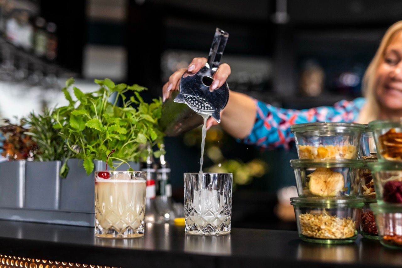 Eine Barkeeperin gießt einen Cocktail durch ein Barsieb in ein Glas mit Eis, umgeben von Kräutern und Zutaten für Getränke.