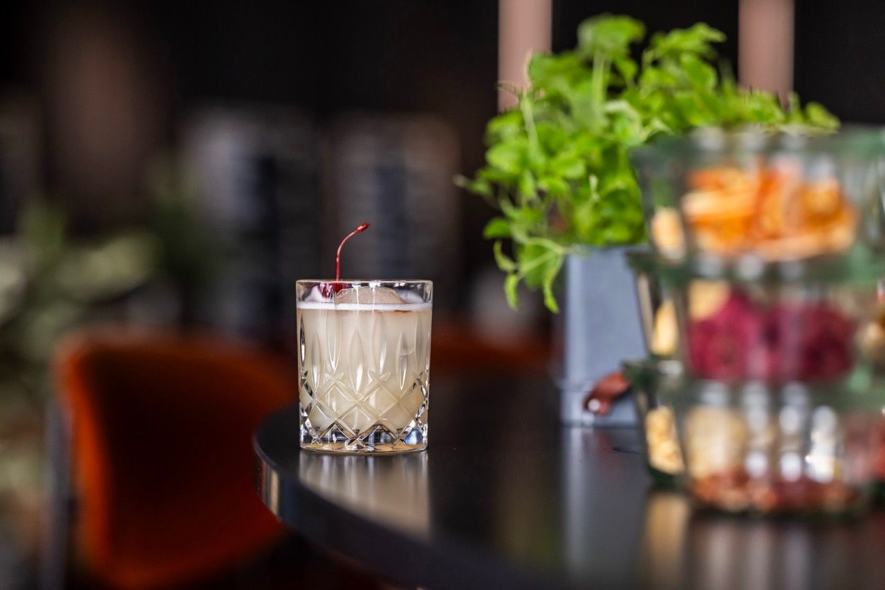Ein Cocktailglas mit cremigem Getränk und Kirsche steht auf einer dunklen Bar-Theke, im Hintergrund unscharfe Pflanzen und warme, gemütliche Lichtstimmung.