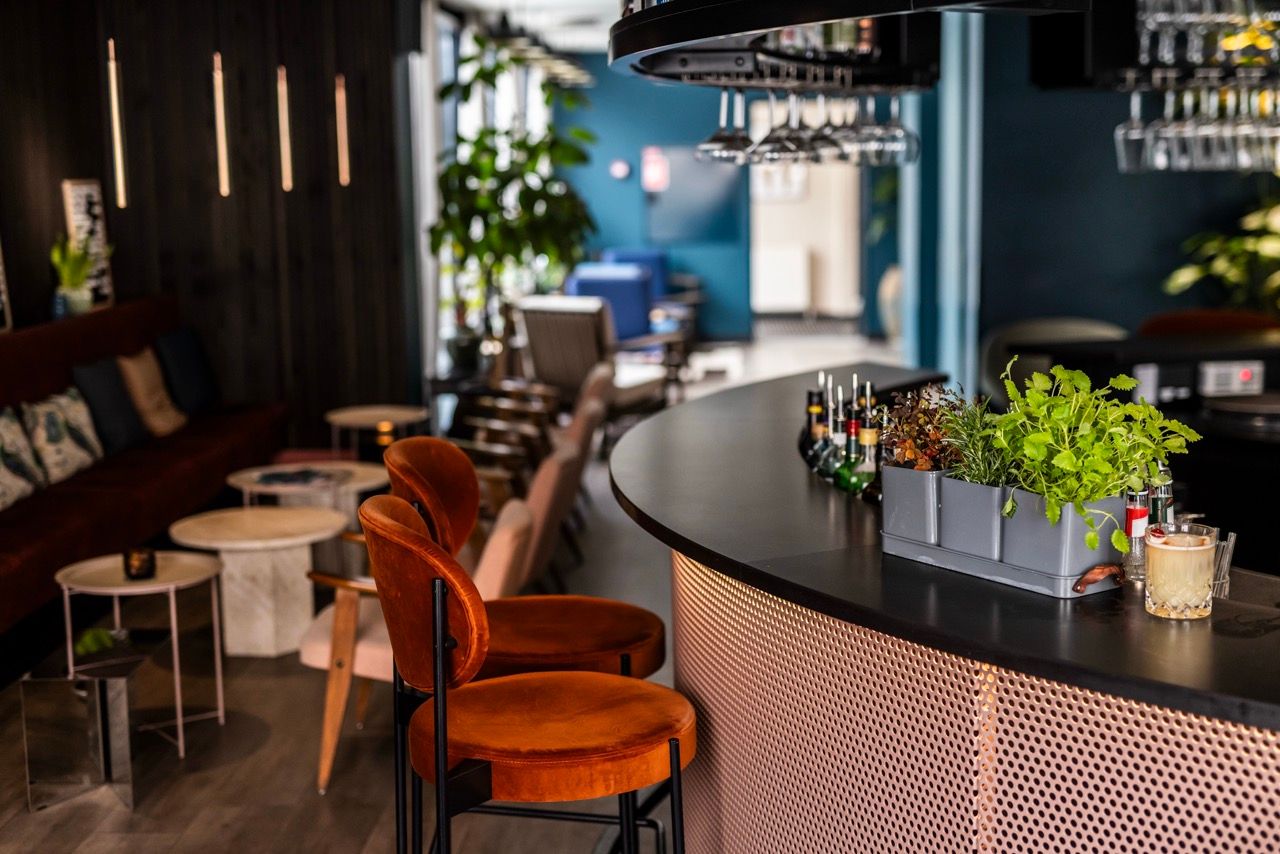 Moderner Hotelbarbereich mit stilvollen roten Barhockern, beleuchteter Theke und Pflanzen, der eine einladende Atmosphäre zum Entspannen und Genießen von Getränken bietet.