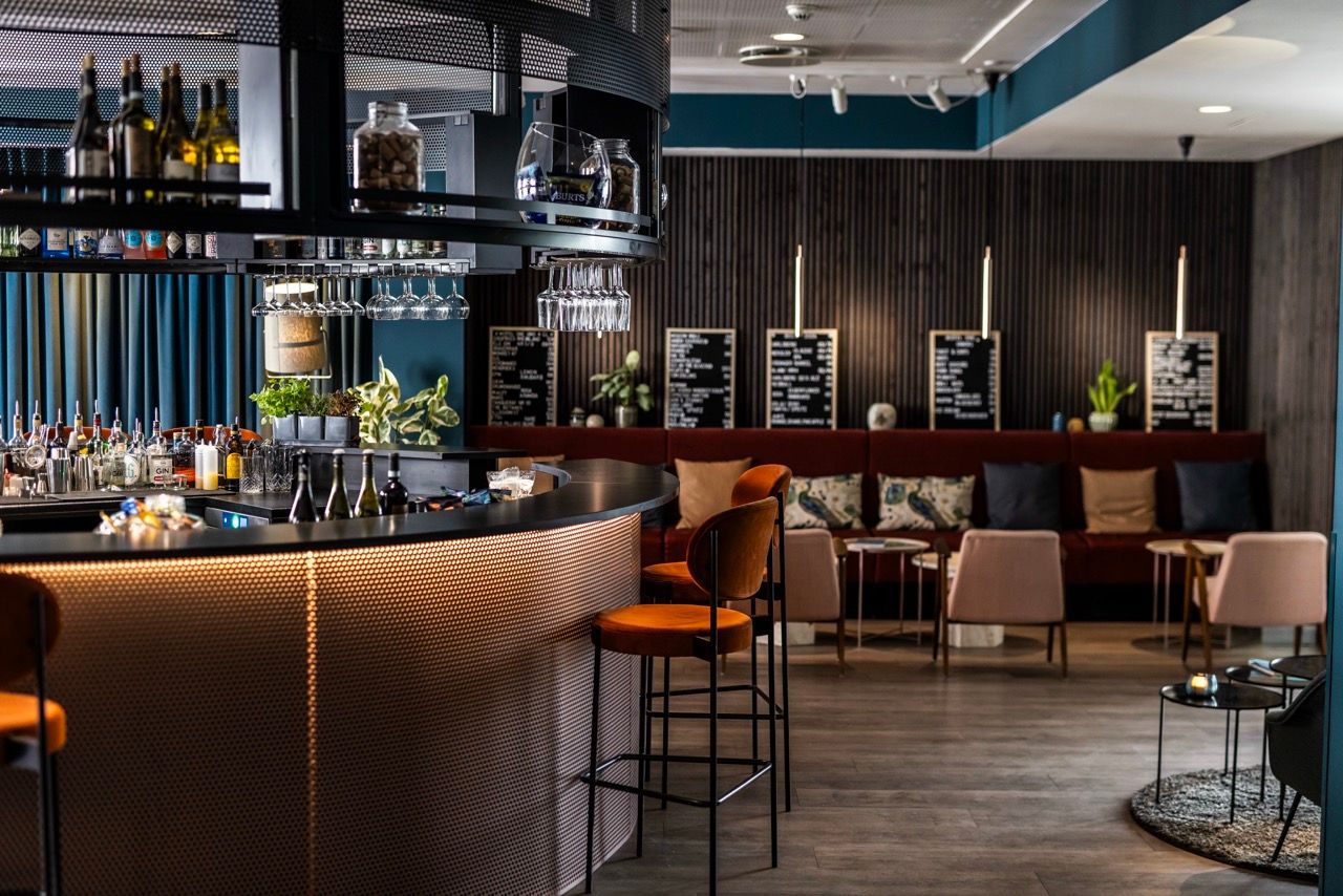 Zeitgemäße Hotelbar mit stilvoll beleuchtetem Tresen, bequemen Barhockern und gemütlicher Sitzecke mit Kissen, die zum Entspannen und Genießen von Getränken einlädt. Modernes, warmes Ambiente für gesellige Abende oder entspannte Momente.