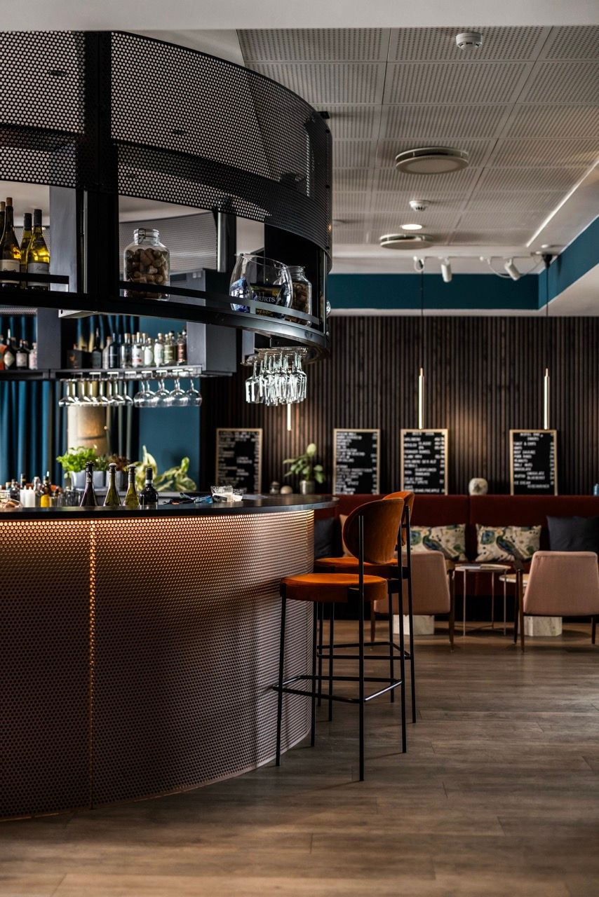 Moderner Hotelbarbereich mit geschwungener Theke, hohen Barhockern und stilvoll arrangierten Getränken. Im Hintergrund gemütliche Sitzgelegenheiten und beleuchtete Tafeln mit Speiseangeboten, die ein einladendes Ambiente für entspannte Abende schaffen.