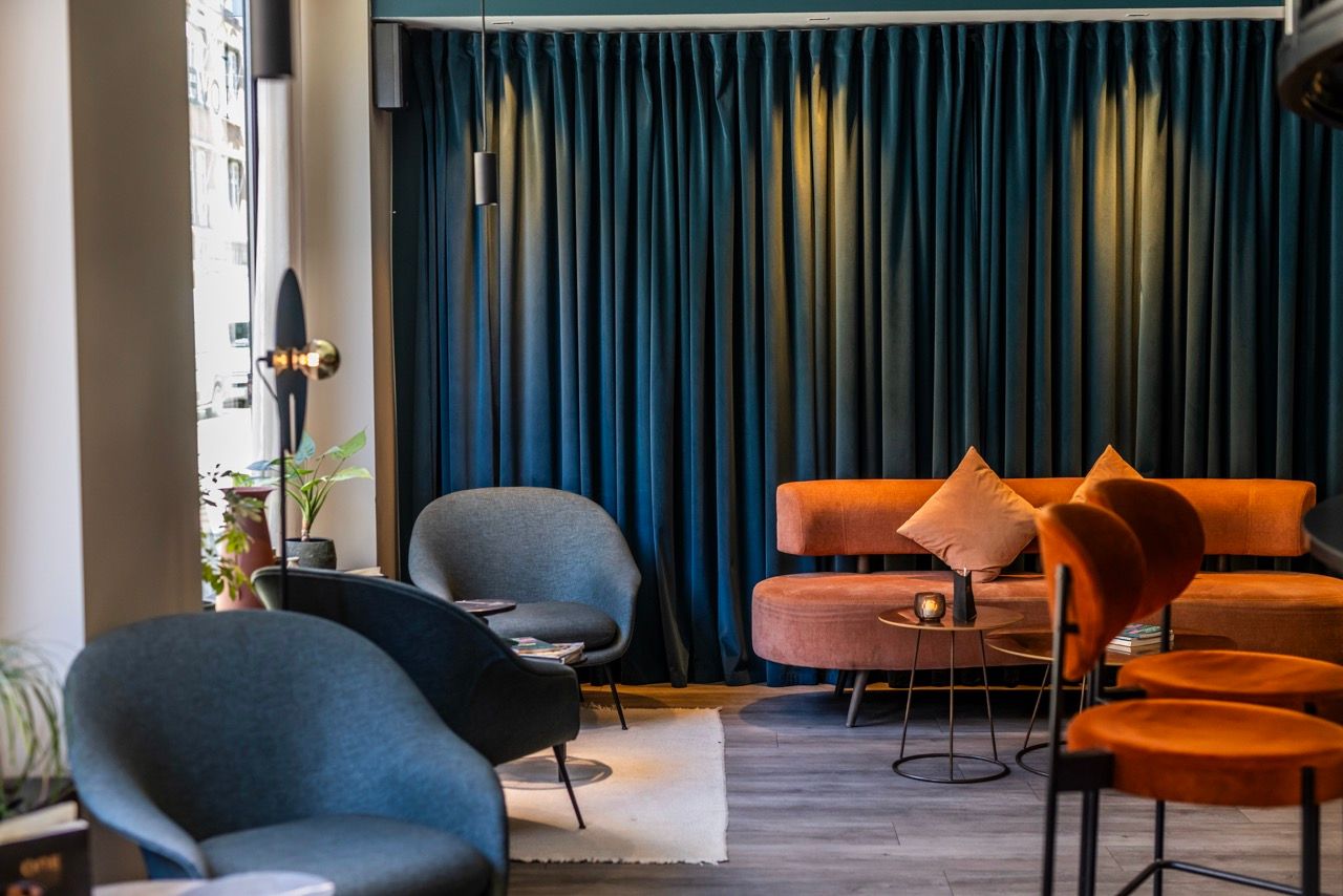 Gemütliche Lounge mit dunkelblauen Vorhängen, zwei blauen Sesseln, einem orangefarbenen Sofa und passenden Stühlen. Sanfte Beleuchtung sorgt für entspannte Atmosphäre, ideal zum Verweilen oder Treffen.