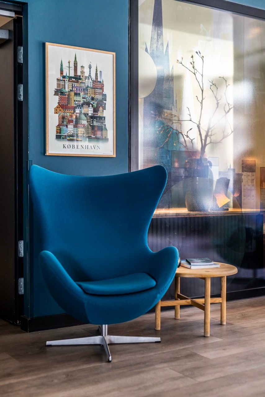 Moderner, dunkelblauer Lounge-Sessel vor dunkelblauer Wand mit stilisiertem Stadtbild von Kopenhagen. Daneben kleiner Holztisch mit Büchern, im Hintergrund Glaswand mit Dekoration und Pflanzen. Ruhige und einladende Atmosphäre im Warte- oder Entspannungsbereich.