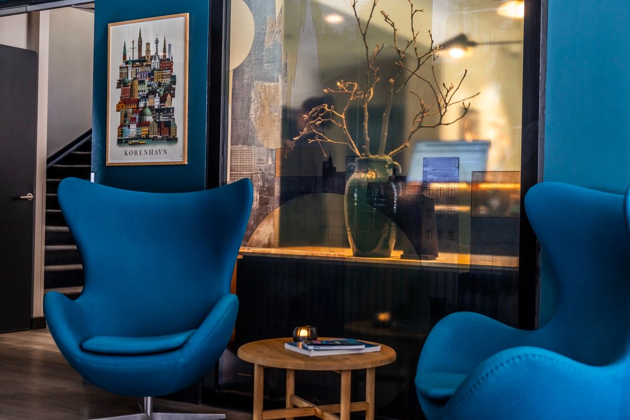 Moderne Lounge-Ecke mit zwei komfortablen blauen Sesseln, einem runden Holztisch und dekorativem Pflanzenarrangement in Vase, stilvoll eingerahmt von künstlerischer Wandgestaltung.