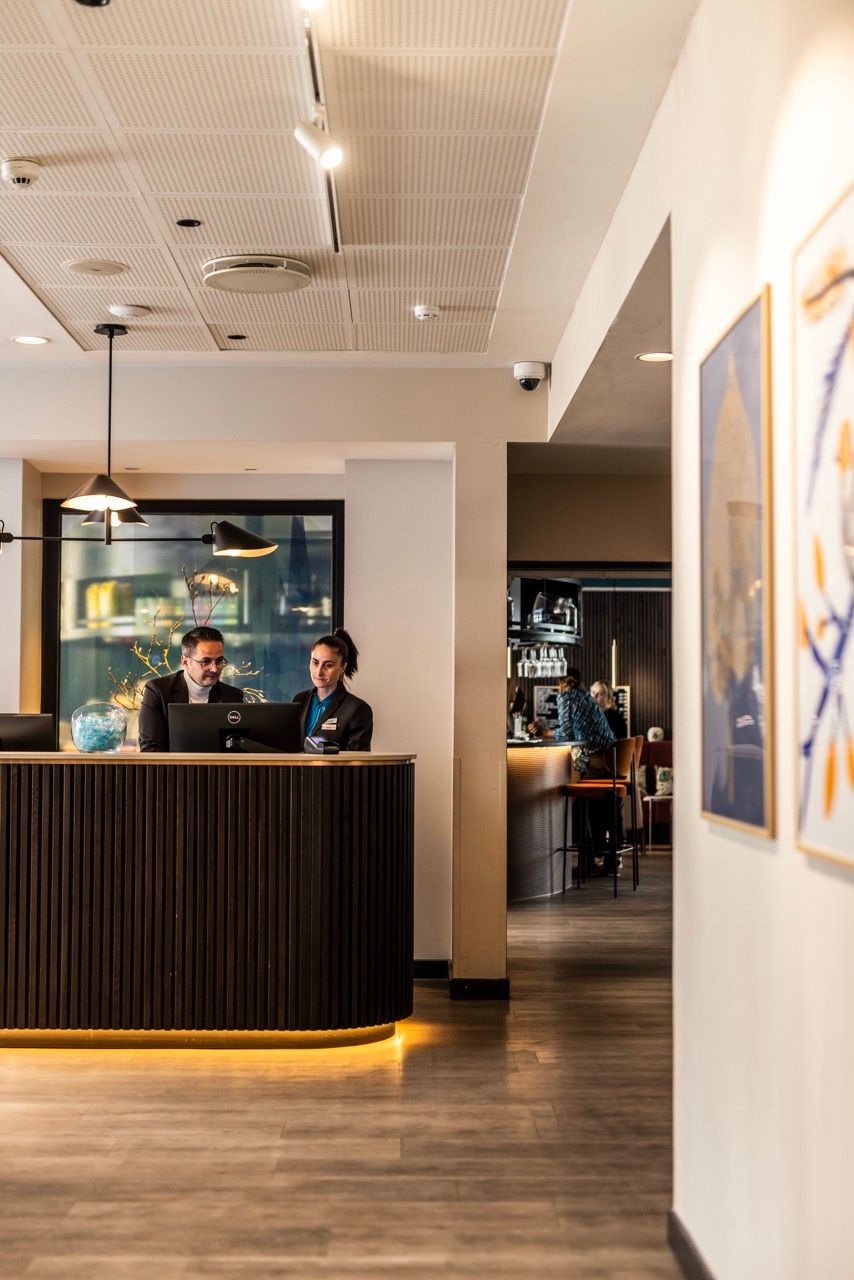 Zwei Mitarbeiter an der Hotelrezeption beraten Gäste, moderner Empfangsbereich mit warmem Licht und sachlicher Atmosphäre.