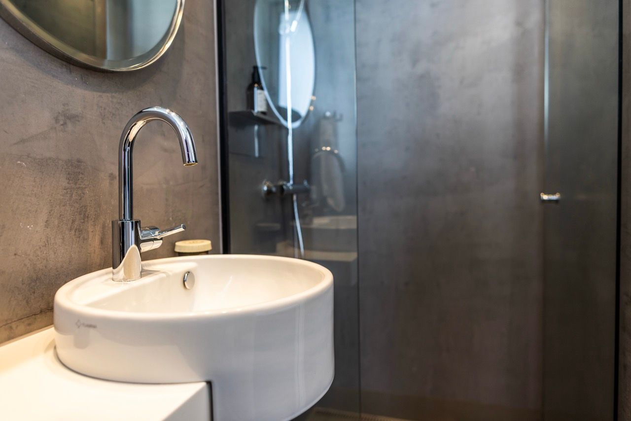 Moderner Hotelbadezimmer-Waschbecken mit elegantem Wasserhahn, rundem Spiegel und begehbarer Dusche mit Glaswand, in schlichter, stilvoller Gestaltung.