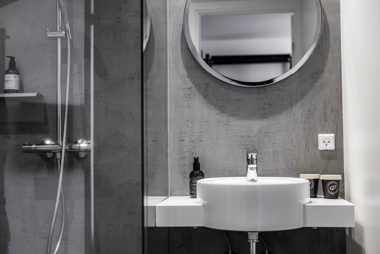 Modernes Badezimmer mit runder Spiegelbeleuchtung, weißem Waschbecken und Glas-Duschkabine, das eine schlichte und saubere Atmosphäre für einen komfortablen Aufenthalt schafft.