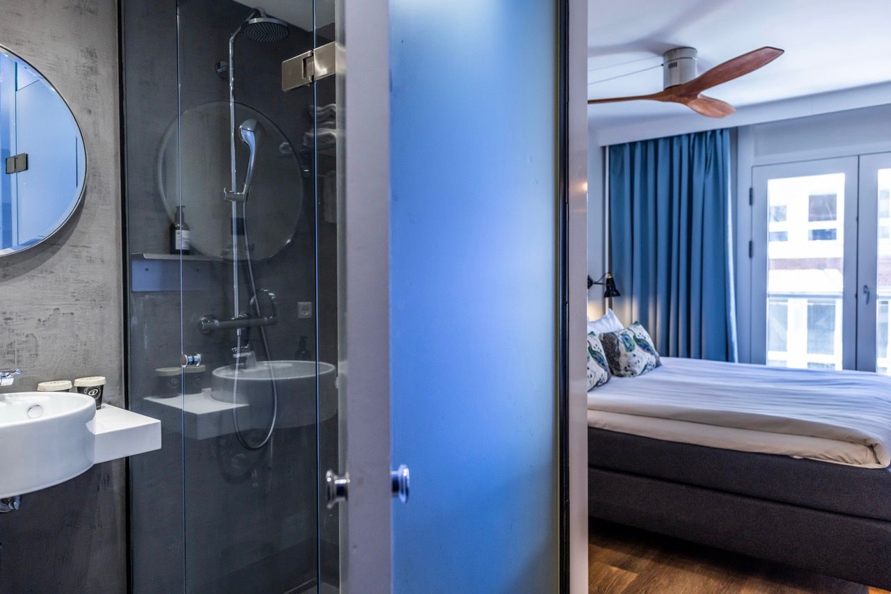 Modernes Hotelzimmer mit großem Bett, blauen Vorhängen und Deckenventilator, angrenzendem Badezimmer mit Glasdusche und rundem Spiegel. Helles, stilvolles Ambiente, geeignet für komfortablen Aufenthalt.