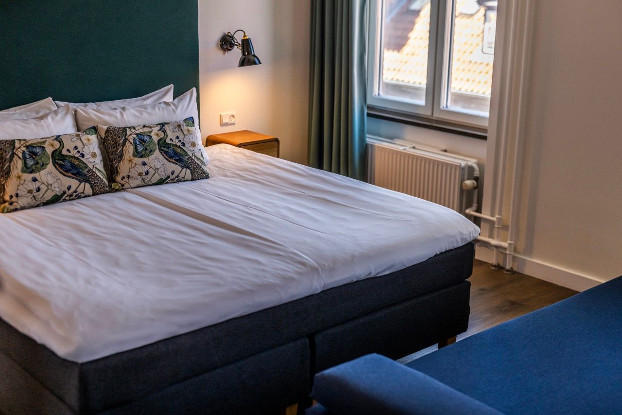 Hell eingerichtetes Hotelzimmer mit großem, bequemem Doppelbett, dekorativen Kissen und großem Fenster, das Tageslicht hereinlässt. Ruhige, gemütliche Atmosphäre für erholsamen Schlaf.