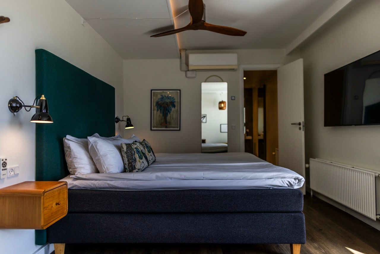 Hell eingerichtetes Schlafzimmer mit großem Doppelbett, bequemen Kopfkissen, dunkelgrünem Kopfteiler, Wandleuchten, Ventilator an der Decke und modernem Flachbildschirm, das Ruhe und Komfort für erholsamen Schlaf verspricht.