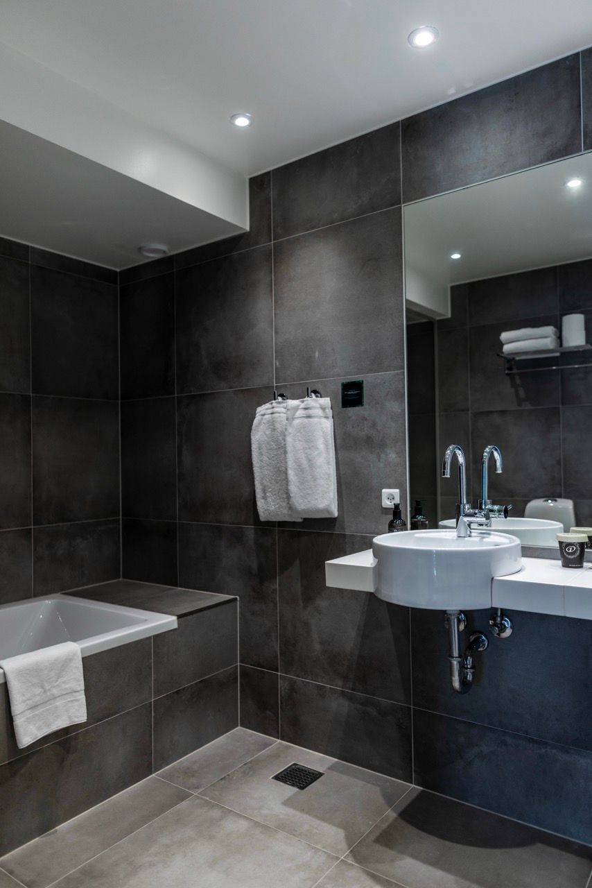 Modernes Badezimmer mit großer Badewanne, weißem Waschbecken, dunklen Fliesen und Handtüchern, das ein elegantes und gepflegtes Ambiente vermittelt.
