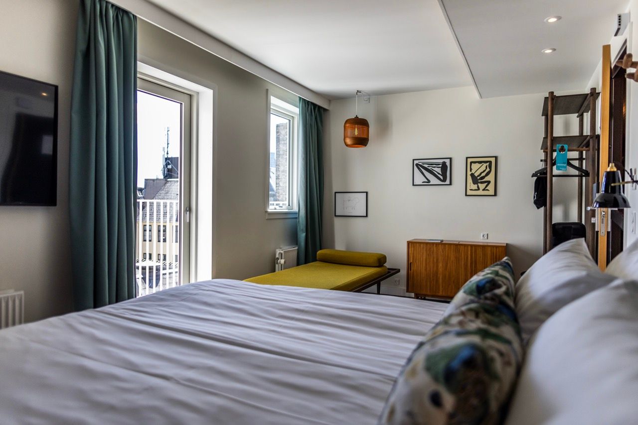 Modernes Hotelzimmer mit großem, bequemem Bett, Tagesbett am Fenster, zurückhaltender, eleganter Einrichtung und natürlichem Tageslicht. Ruhige Atmosphäre für erholsamen Aufenthalt in urbaner Umgebung.