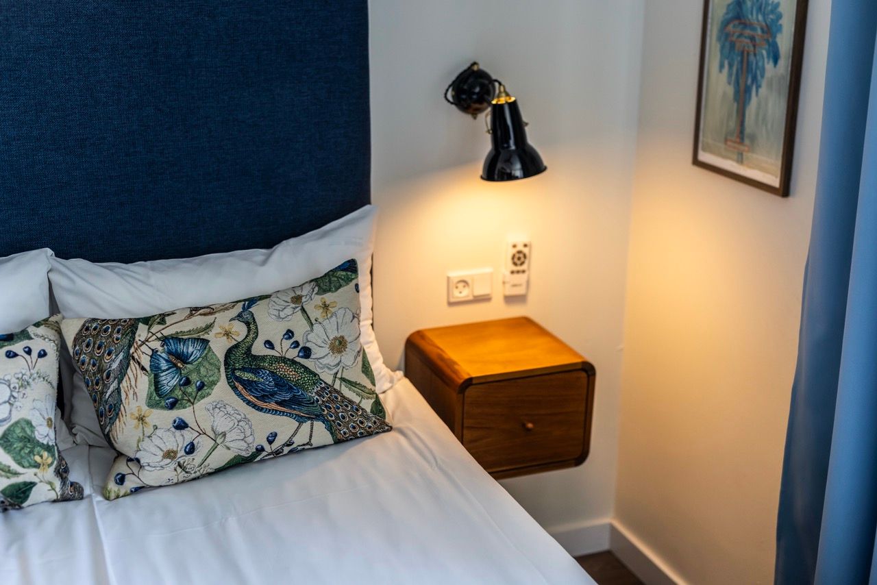 Gemütliche Hotelzimmer-Ecke mit weißem Bett, dekorativen Kissen mit Pfauenmotiv, dunkelblauem Kopfteil und einer warmen Wandlampe über einem kleinen, hölzernen Nachttisch.