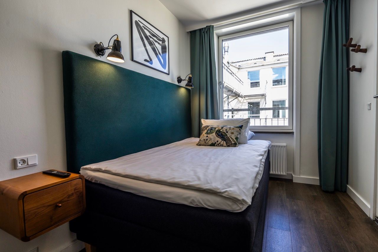 Kompaktes Hotelzimmer mit großem Fenster, gemütlichem Doppelbett mit dunklem Kopfteil, Nachttisch und moderner Beleuchtung, hell und einladend für erholsamen Schlaf in zentraler Lage.
