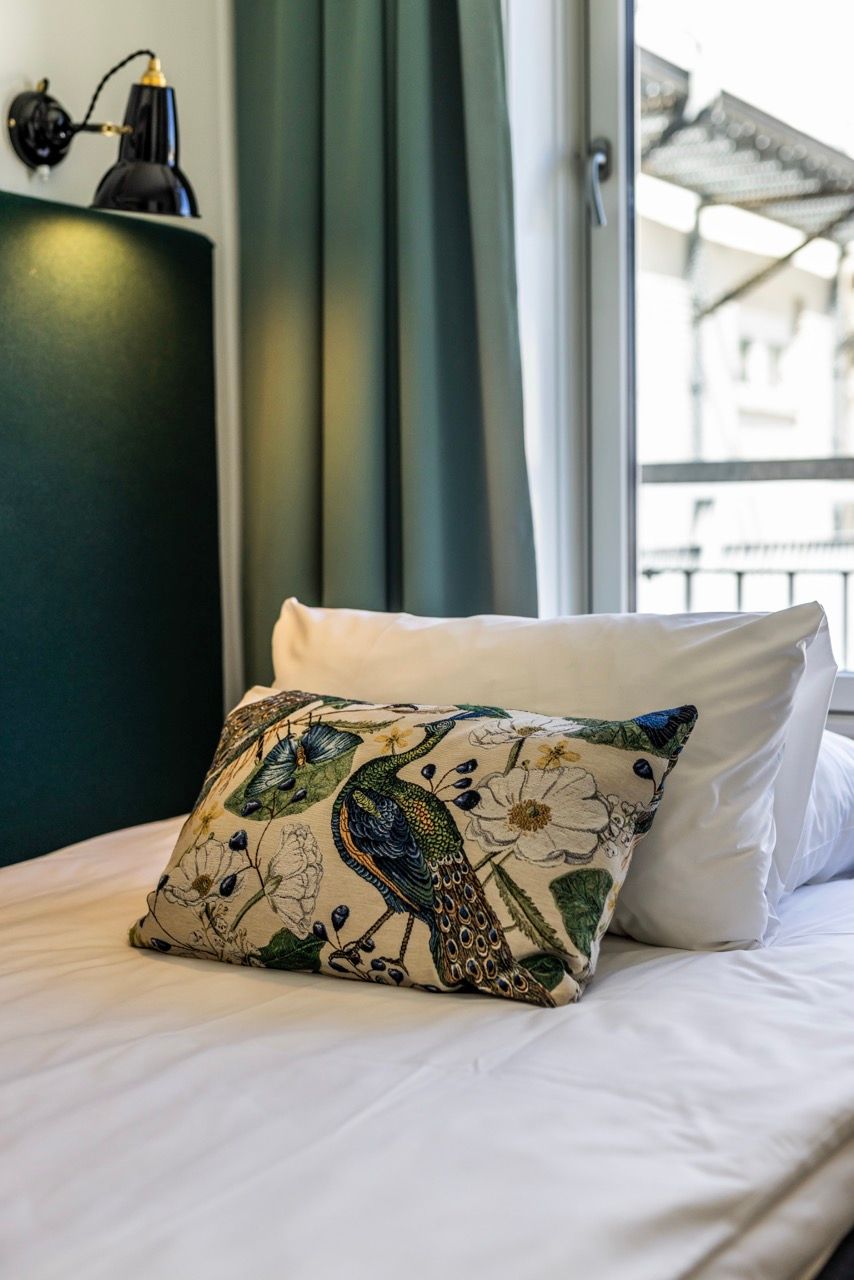 Gemütliches Einzelbett mit weißer Bettwäsche und dekorativem Kissen mit Pfauenmotiv, neben einem Fenster mit Blick auf einen Balkon. Sanfte Beleuchtung und grüne Vorhänge schaffen eine ruhige Atmosphäre.