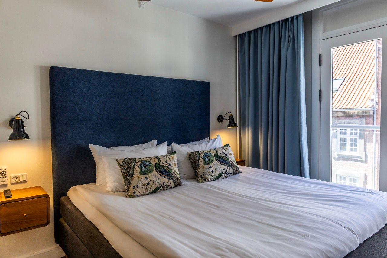 Gemütliches Doppelbett mit dunkelblauem Kopfteil, weißen Bettlaken und dekorativen Kissen, sanfte Beleuchtung, angrenzender Balkon mit Blick auf Nachbargebäude, ruhige und helle Atmosphäre.