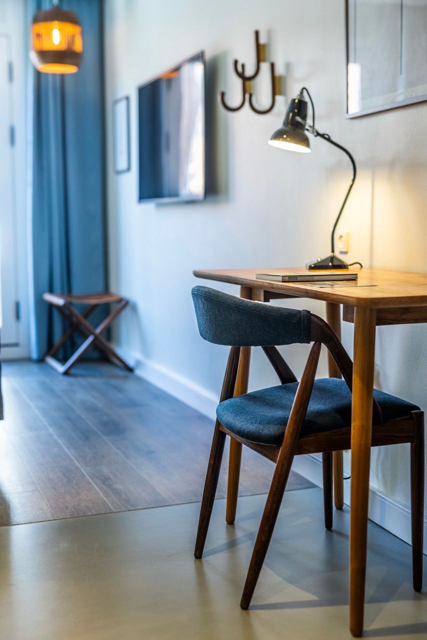 Moderner Arbeitsbereich mit Holzschreibtisch, bequemen Polsterstuhl und Schreibtischlampe in einem hellen Hotelzimmer mit großem Fenster und stilvoller Dekoration.