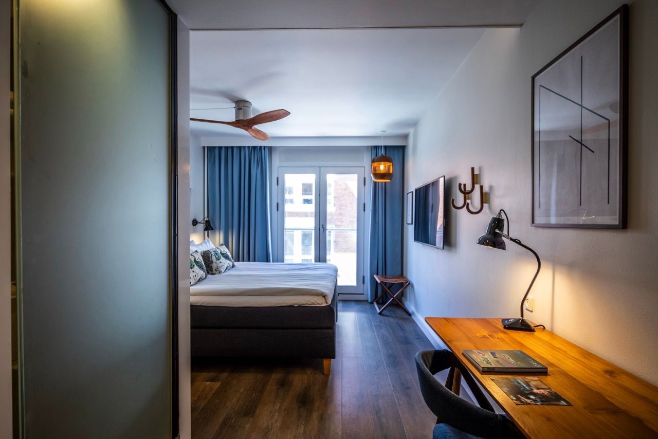Modern eingerichtetes Hotelzimmer mit großem Doppelbett, Schreibtisch, Fernseher und eigenem Balkon. Helle, freundliche Atmosphäre mit Holzfußboden, blauen Vorhängen und stilvollen Akzenten. Ideal für Geschäftsreisende und Urlauber.