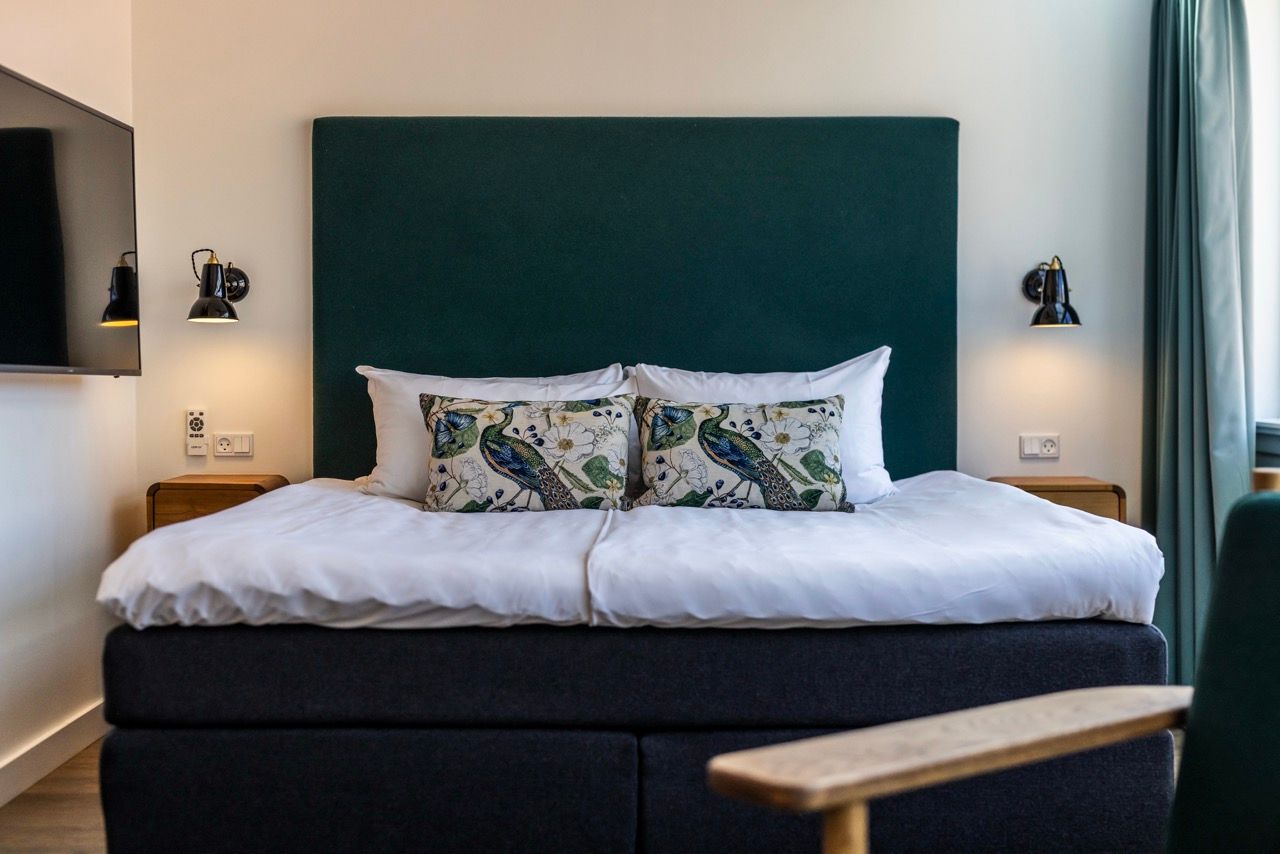 Modernes Hotelzimmer mit großem Doppelbett, dunklem Kopfteil und dekorativen Kissen mit Pfauenmotiven. Angenehme Beleuchtung durch Leselampen an beiden Seiten, ruhige und einladende Atmosphäre für erholsamen Schlaf.