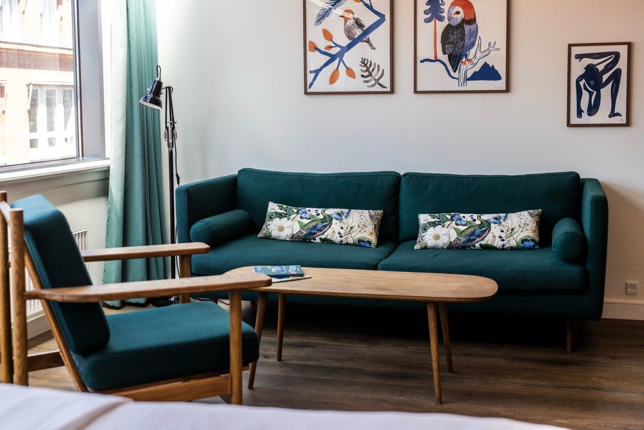Gemütliche Sitzecke mit dunkelgrünem Sofa und passendem Sessel, hellem Holztisch und modernen Tiermotiven an der Wand, lichtdurchflutet durch ein großes Fenster mit Vorhang.