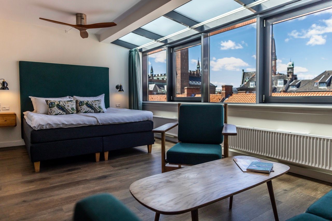 Modern eingerichtetes Hotelzimmer mit großem Doppelbett, gemütlichem Sitzbereich und Panoramafenstern, die einen Blick auf historische Stadtgebäude bieten. Natürliche Lichtquelle durch Oberlichter und Fenster sorgt für helle, freundliche Atmosphäre.