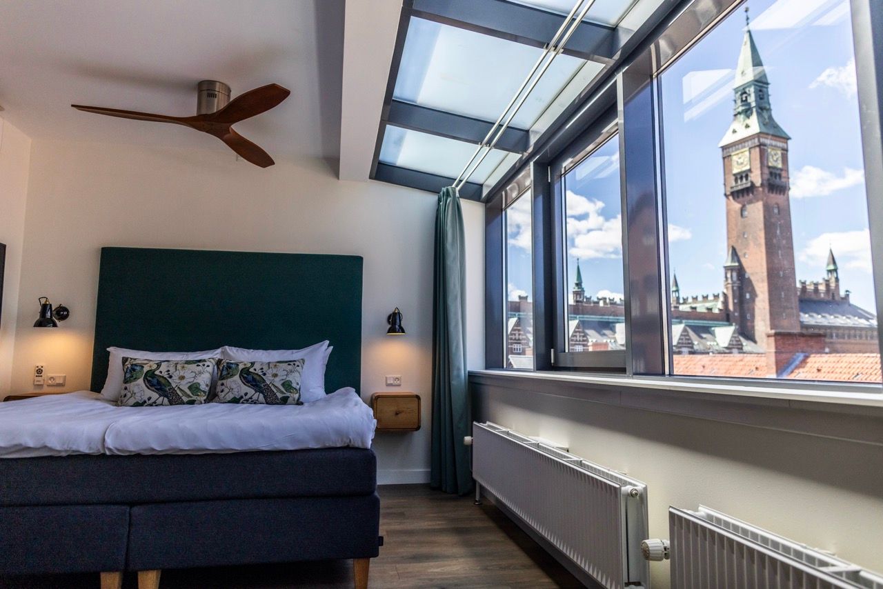 Helles Hotelzimmer mit großem Fenster und Glasdach, Blick auf historischen Turm. Modernes, gemütliches Doppelbett, klare Linien, ruhige Atmosphäre, ideal für erholsamen Aufenthalt in der Stadt.