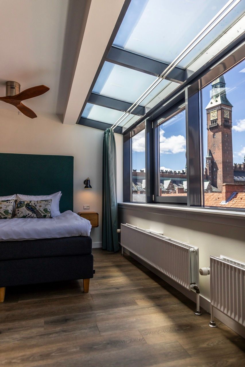 Helles, modern eingerichtetes Hotelzimmer mit großem Panoramafenster und Glasdach, das einen klaren Blick auf einen historischen Uhrturm und die Stadt bietet. Gemütliches Bett und warme Holzböden schaffen ein einladendes Ambiente.