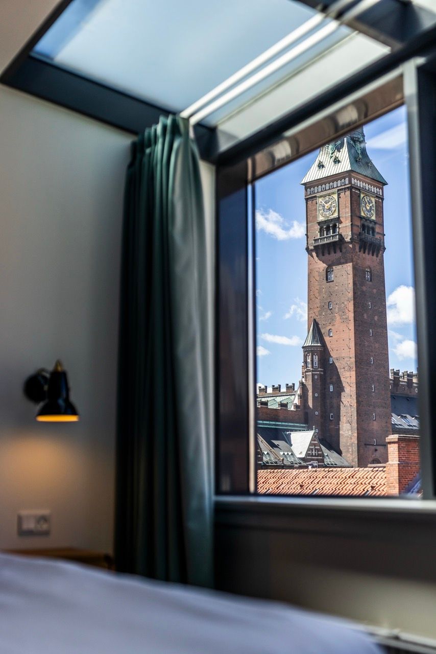 Blick aus einem Hotelzimmerfenster auf einen historischen Uhrturm bei klarem Himmel, sanftes Tageslicht durch das Fenster, gemütliche Atmosphäre im Zimmer mit Vorhang und Wandlampe.