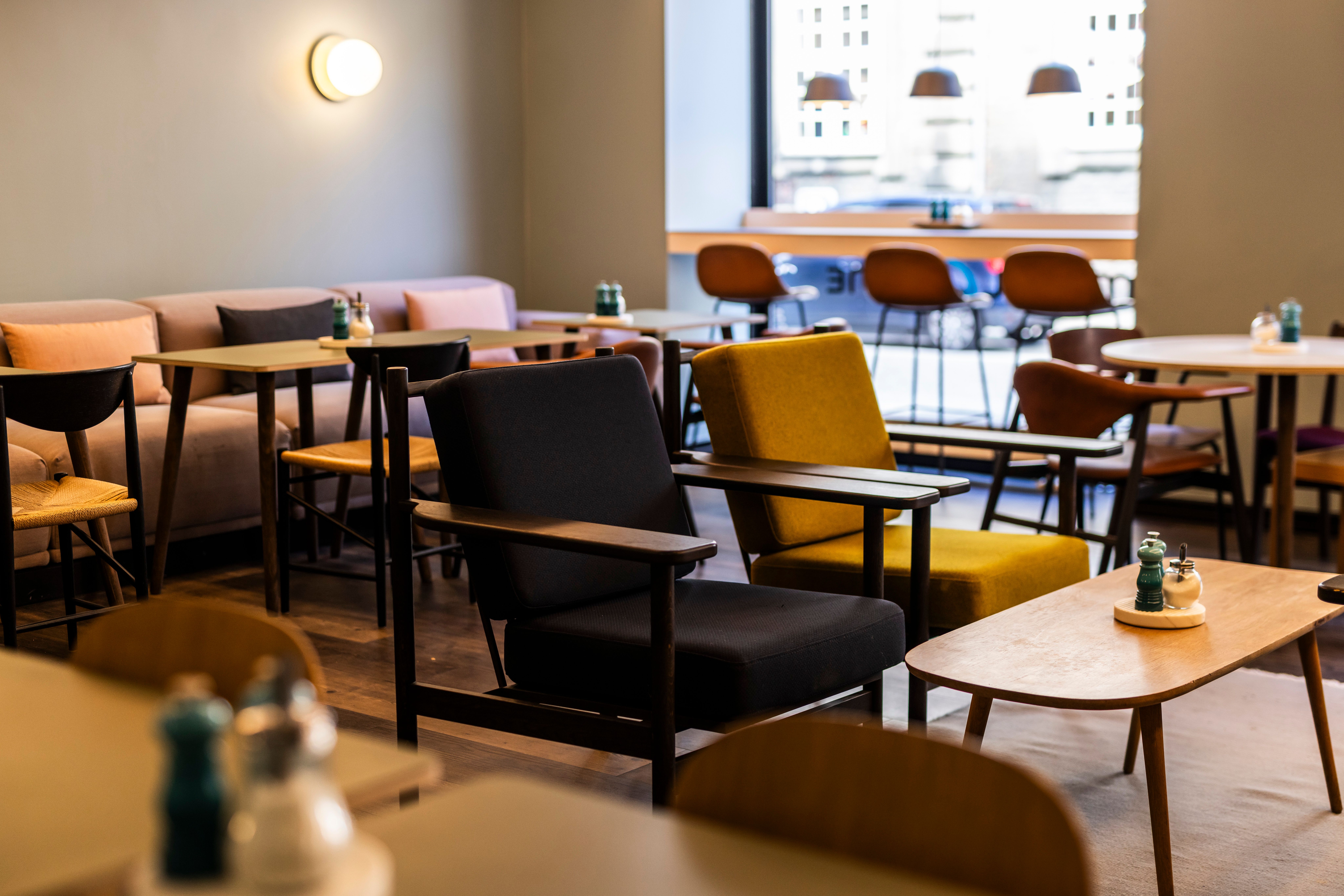 Gemütlicher Lounge-Bereich mit modernen Sesseln und Holztischen in warmem, einladendem Ambiente, ideal zum Entspannen oder für informelle Treffen. Große Fenster sorgen für viel Tageslicht und Blick auf die Stadt.