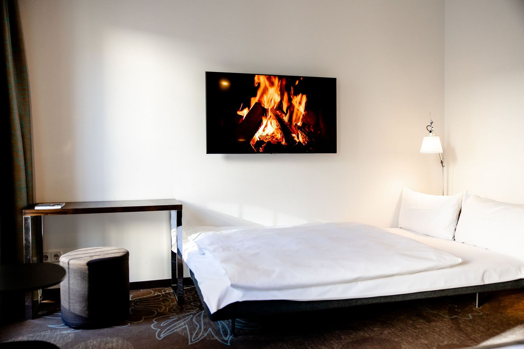 Hotel Motel One Leipzig-Augustusplatz Gemütliches Schlafzimmer mit großem Bett, weicher Beleuchtung und einem Bildschirm, der ein knisterndes Kaminfeuer zeigt. Ruhe und Wohlfühlatmosphäre für erholsamen Aufenthalt.