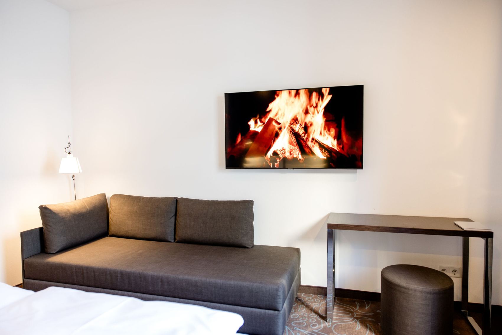 Hotel Motel One Leipzig-Augustusplatz Gemütliche Sitzecke mit dunkelgrauem Sofa, schlichtem Schreibtisch und Hocker, an der Wand ein Fernseher, der ein knisterndes Kaminfeuer zeigt. Helle, ruhige Raumgestaltung für entspannte Atmosphäre.