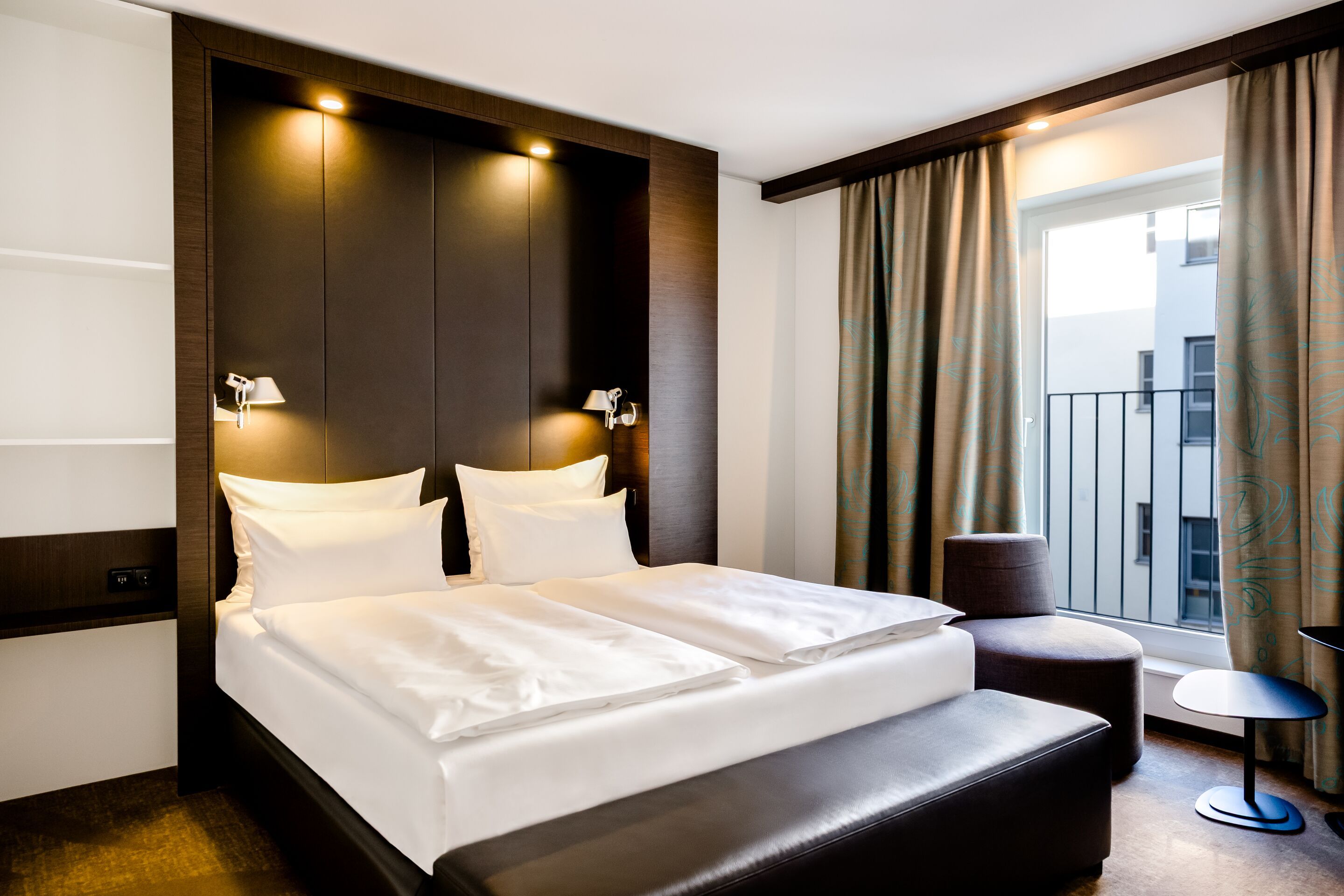 Hotel Leipzig Augustusplatz Motel One Komfortables Hotelzimmer mit großem Doppelbett, weicher Beleuchtung und modernem, dunklem Holzdekor. Sitzbereich mit Sessel und kleinem Tisch neben Fenster mit Blick nach draußen.