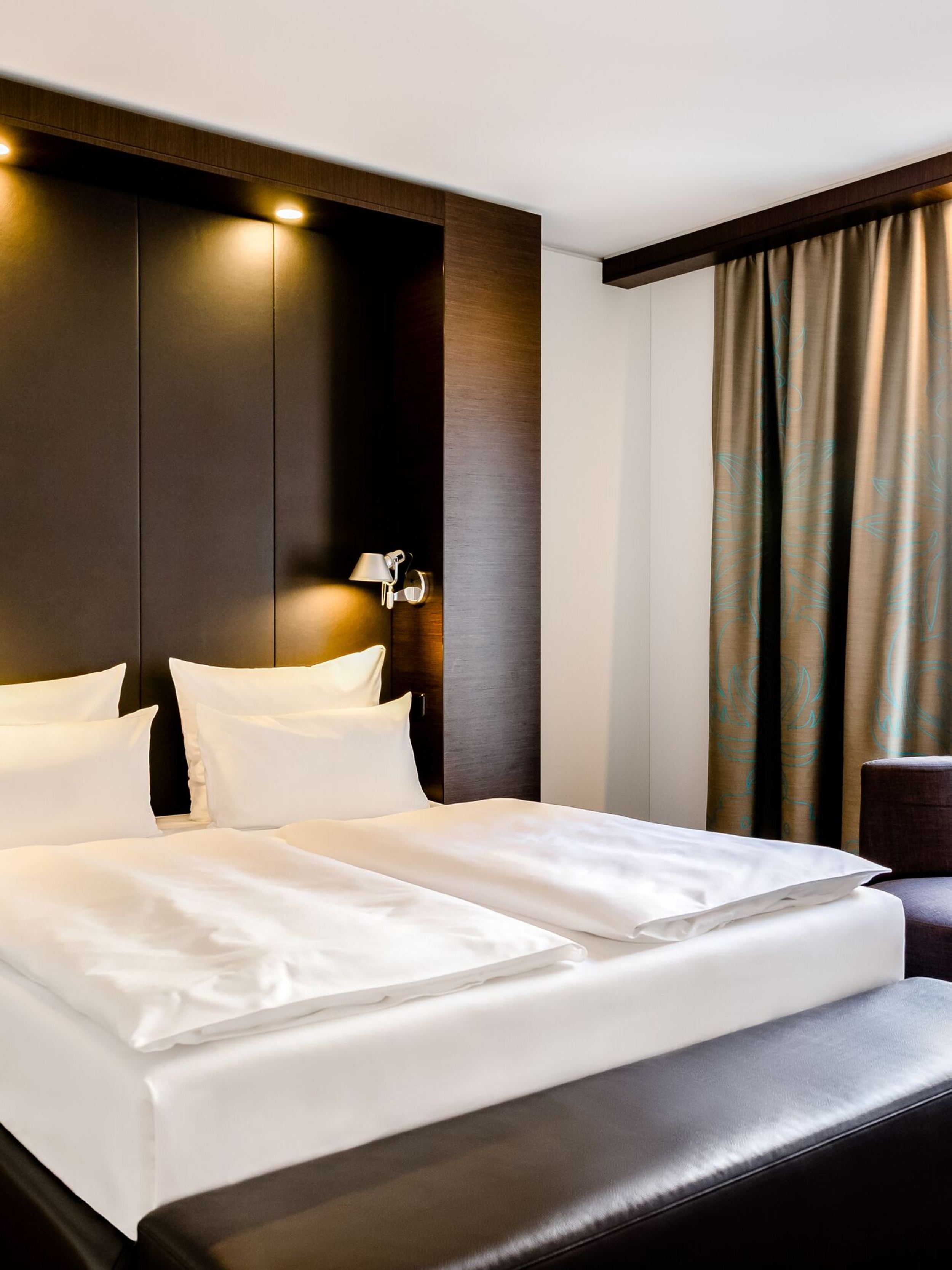 Hotel Leipzig Augustusplatz Motel One Komfortables Hotelzimmer mit großem Doppelbett, weicher Beleuchtung und modernem, dunklem Holzdekor. Sitzbereich mit Sessel und kleinem Tisch neben Fenster mit Blick nach draußen.