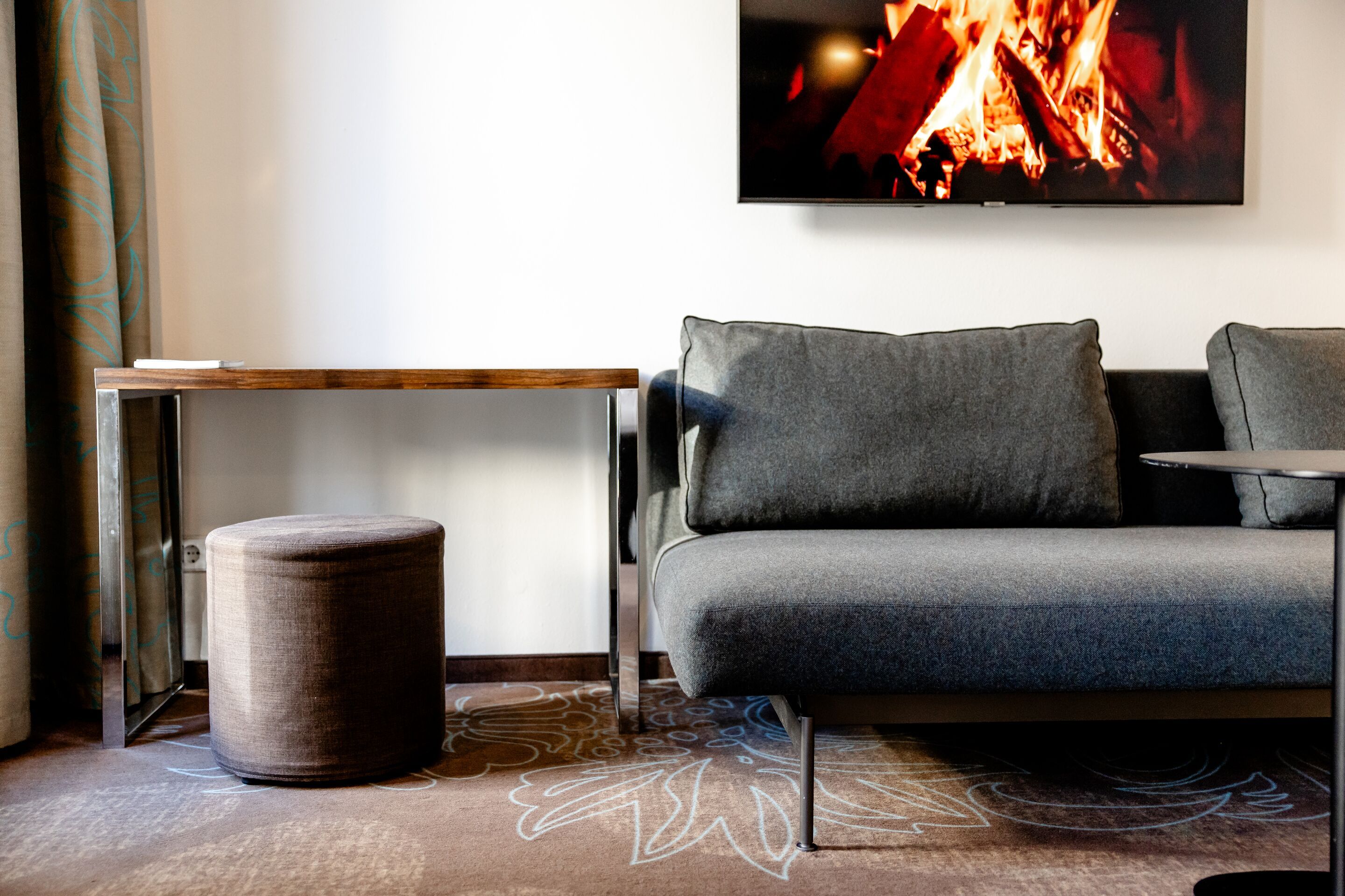 Hotel Leipzig Augustusplatz Motel One moderner Hotelzimmerbereich mit grauem Sofa, kleinem Tisch und gemütlichem Sitzhocker, beleuchtet vom warmen Licht eines digitalen Kamins an der Wand.