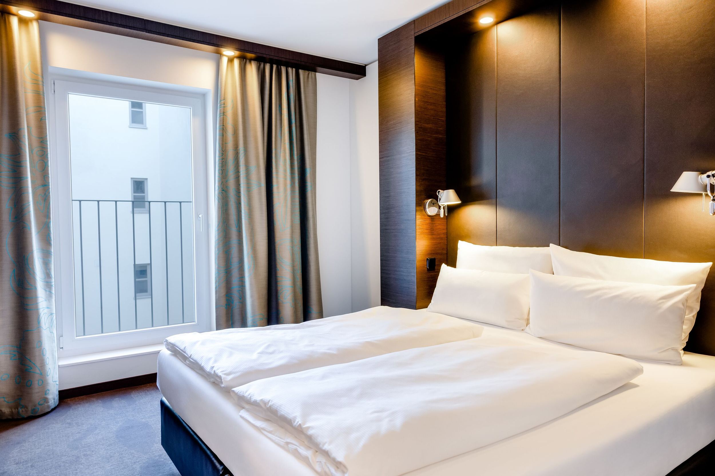 Hotel Leipzig Augustusplatz Motel One Hotelzimmer mit großem Doppelbett, weißen Bettwaren, dunklem Kopfteil und indirekter Beleuchtung. Helles Tageslicht fällt durch ein großes Fenster mit Blick auf Innengebäude. Vorhänge rahmen das Fenster elegant ein.