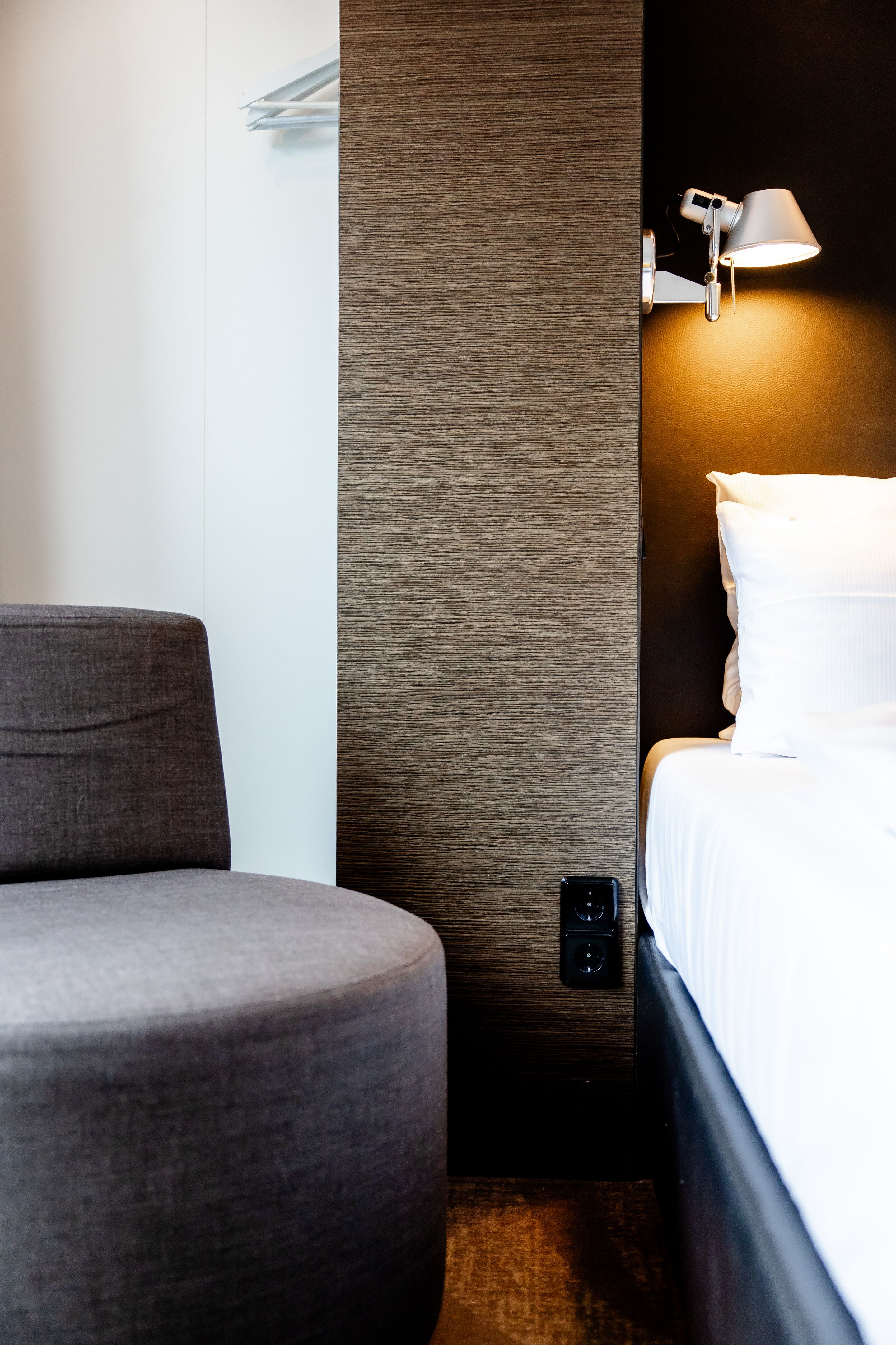 Hotel Leipzig Augustusplatz Motel One halbseitige Ansicht eines modernen Hotelzimmers mit einem grauen Sessel, weißem Bett, Wandlampe und zurückhaltender, eleganter Wandgestaltung.