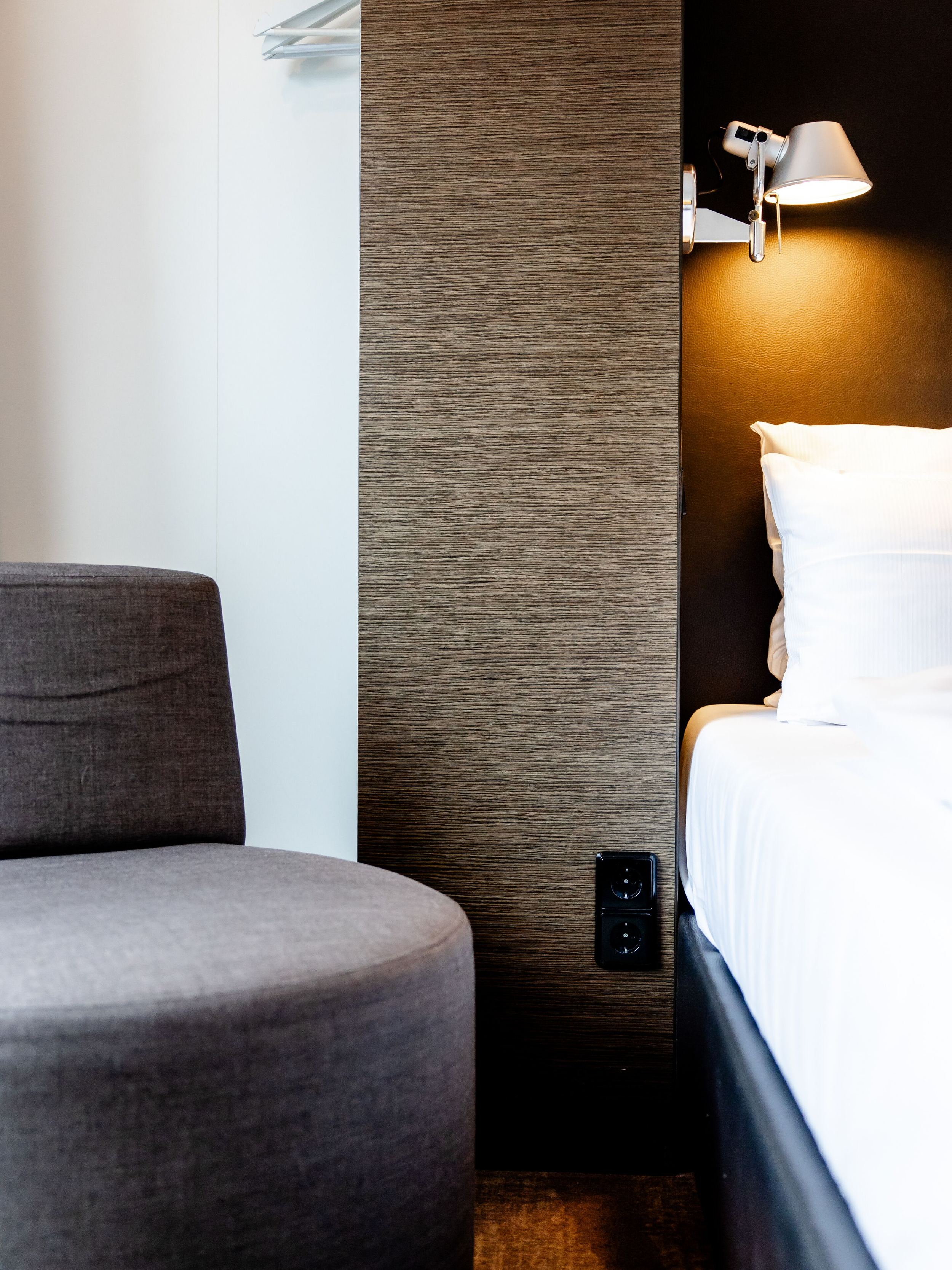 Hotel Leipzig Augustusplatz Motel One halbseitige Ansicht eines modernen Hotelzimmers mit einem grauen Sessel, weißem Bett, Wandlampe und zurückhaltender, eleganter Wandgestaltung.