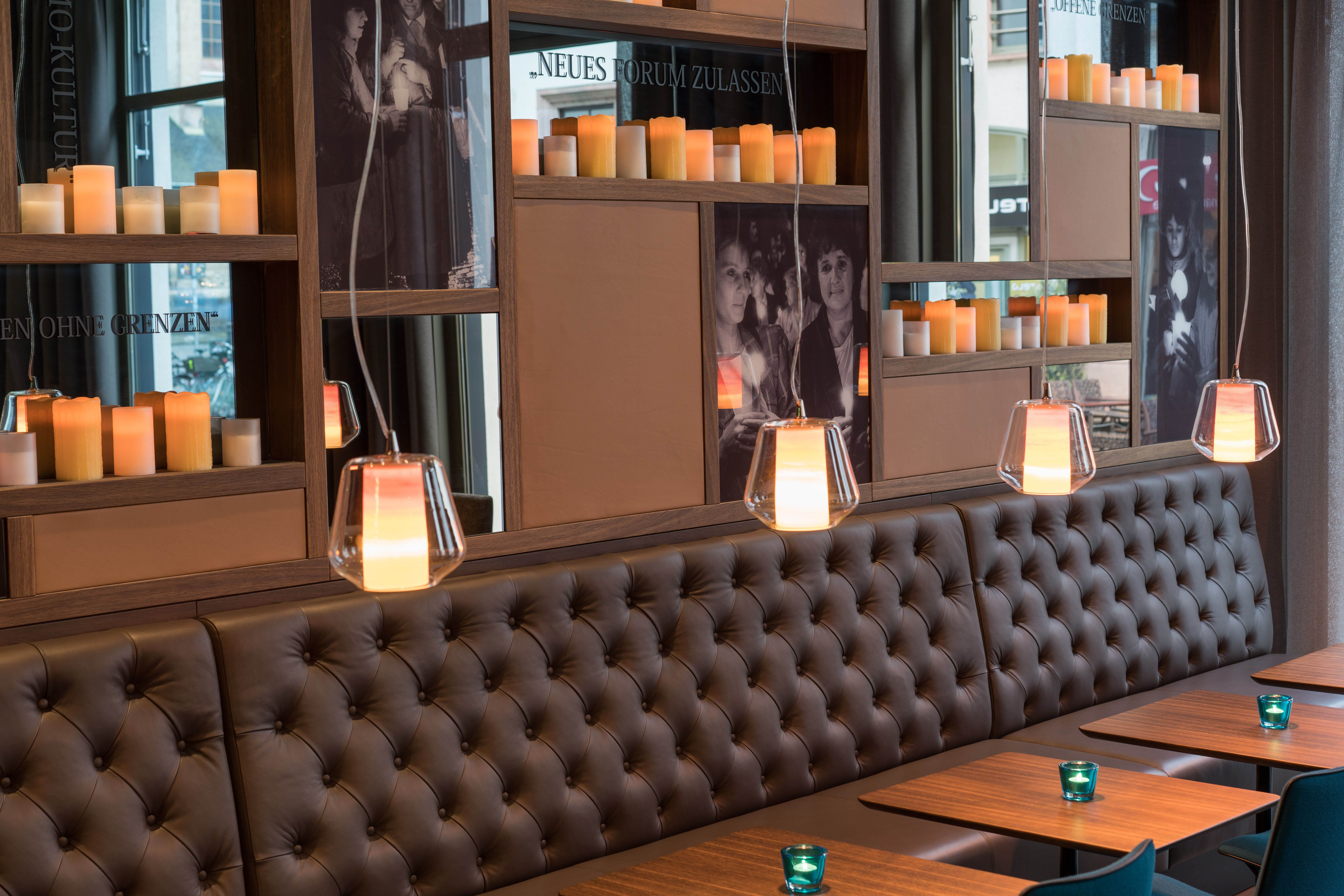 Hotel Leipzig Nikolaikirche Motel One Blick auf Regal mit Kerzen über langer Couch sowie mehrere Tische und Stühle
