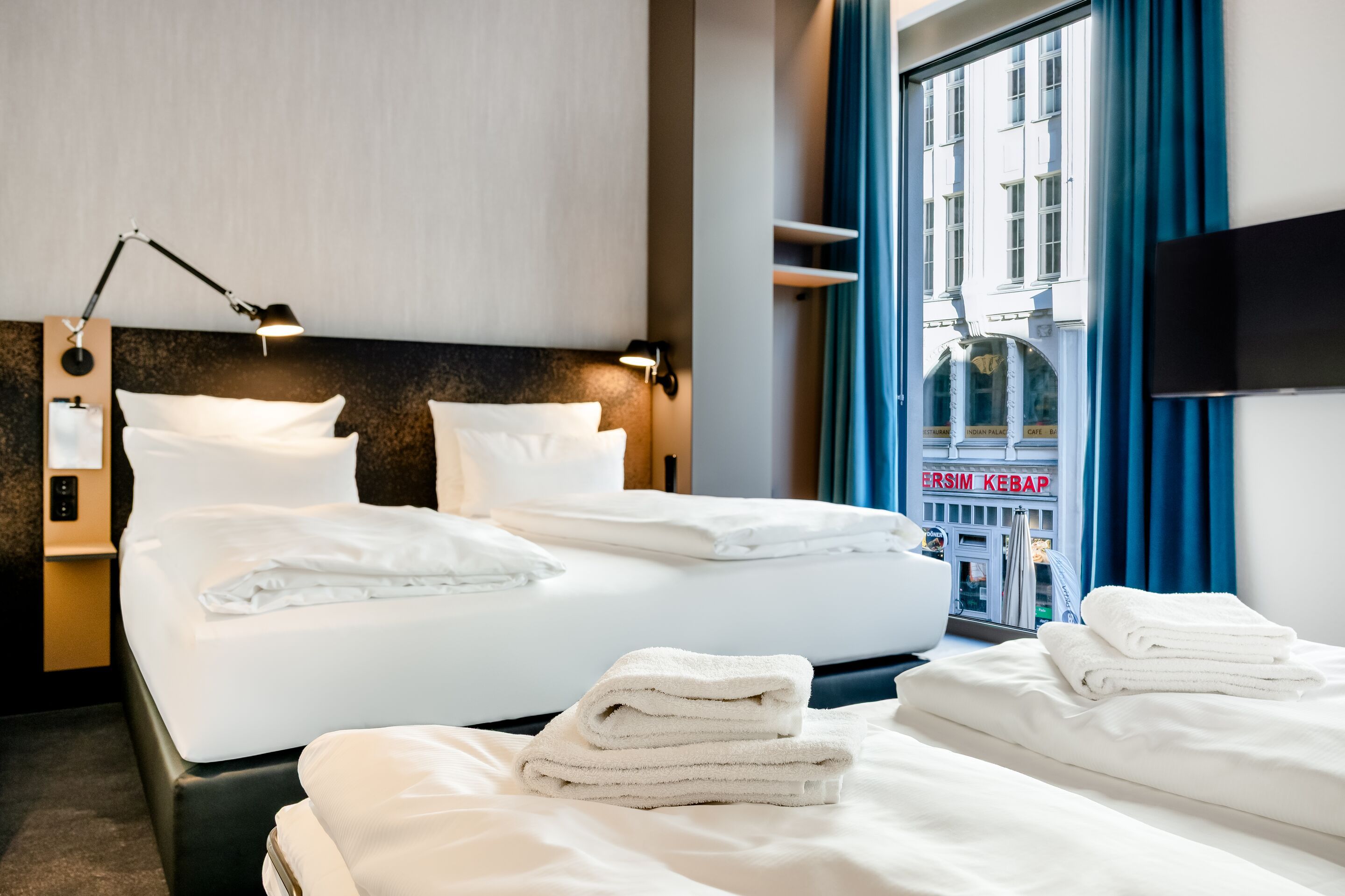 Hell eingerichtetes Hotelzimmer mit zwei Einzelbetten, sauberen weißen Bettwäsche und Handtüchern. Große Fenster mit blauen Vorhängen bieten Stadtblick. Moderne Wandlampen sorgen für angenehmes Licht. Schlichte, komfortable Atmosphäre für erholsamen Aufenthalt.