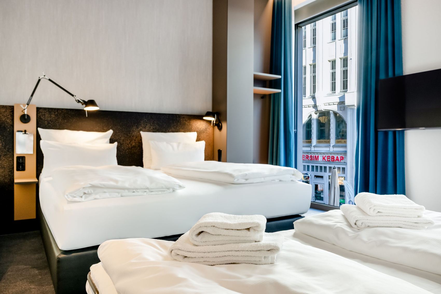 Hell eingerichtetes Hotelzimmer mit zwei Einzelbetten, sauberen weißen Bettwäsche und Handtüchern. Große Fenster mit blauen Vorhängen bieten Stadtblick. Moderne Wandlampen sorgen für angenehmes Licht. Schlichte, komfortable Atmosphäre für erholsamen Aufenthalt.