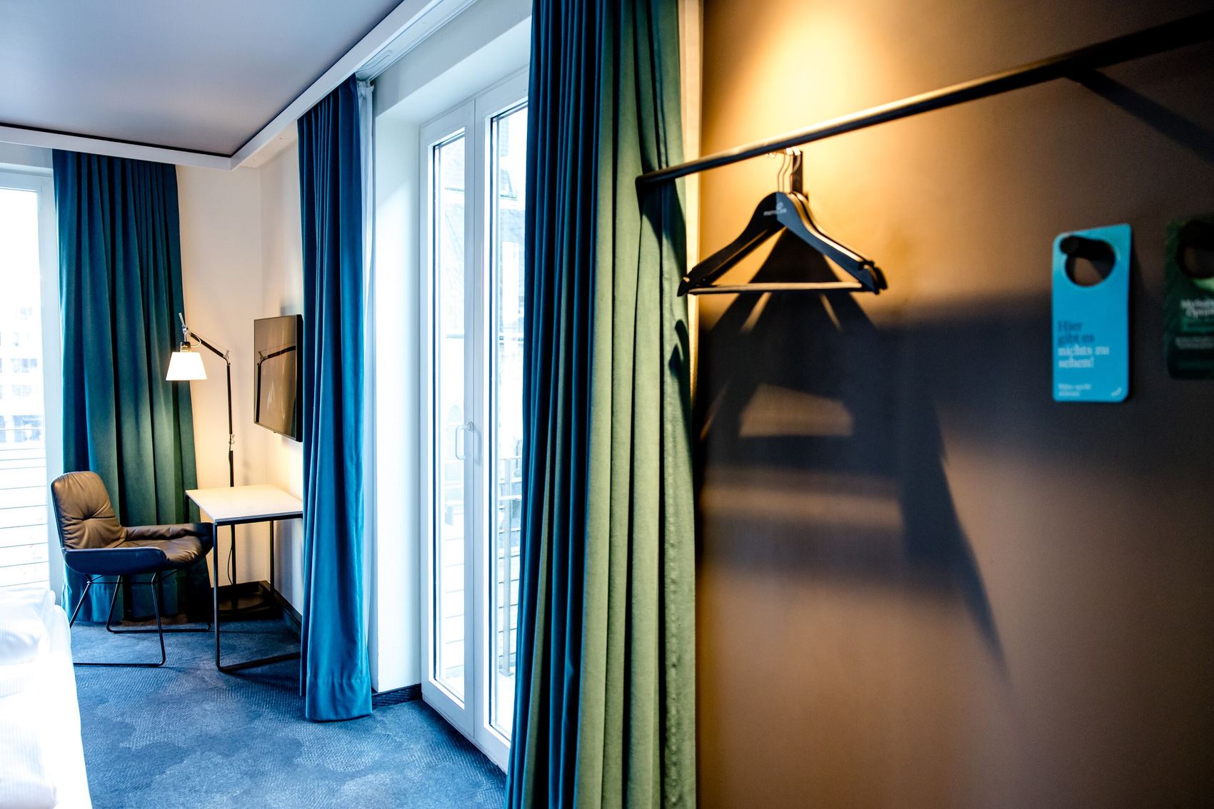 Modernes Hotelzimmer mit Schreibtisch, Sessel und Flachbildfernseher. Große Fenster mit grünen Vorhängen lassen Tageslicht herein. Offene Garderobe an der Wand mit Kleiderbügeln bietet Platz für Kleidung. Behagliche Atmosphäre durch warmes Licht und dezente Farbtöne.